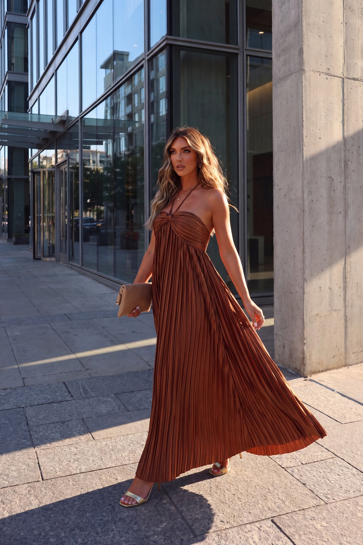 Liora rust satin plisse halter maxi dress, worn walking past a glass fronted building