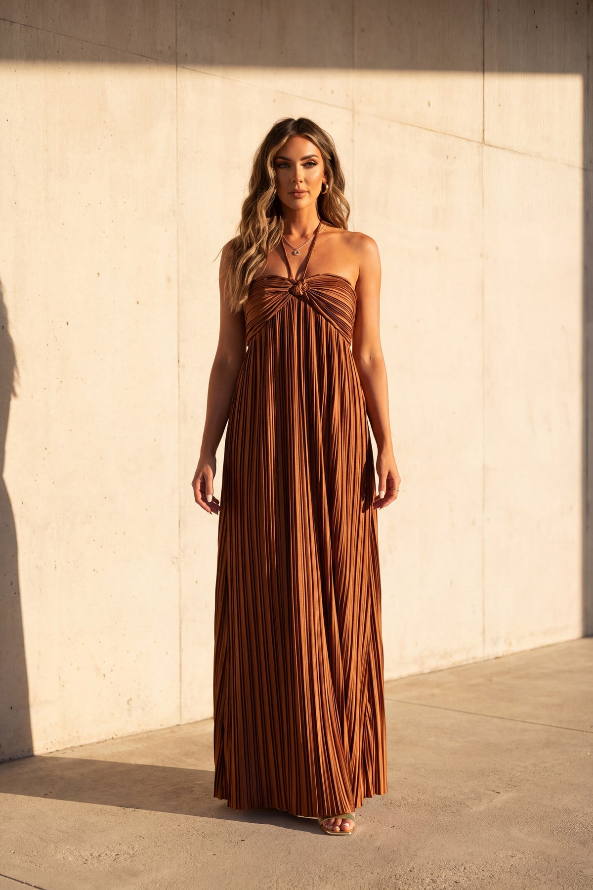 Liora rust satin plisse halter maxi dress, worn full length against a sunlit concrete wall